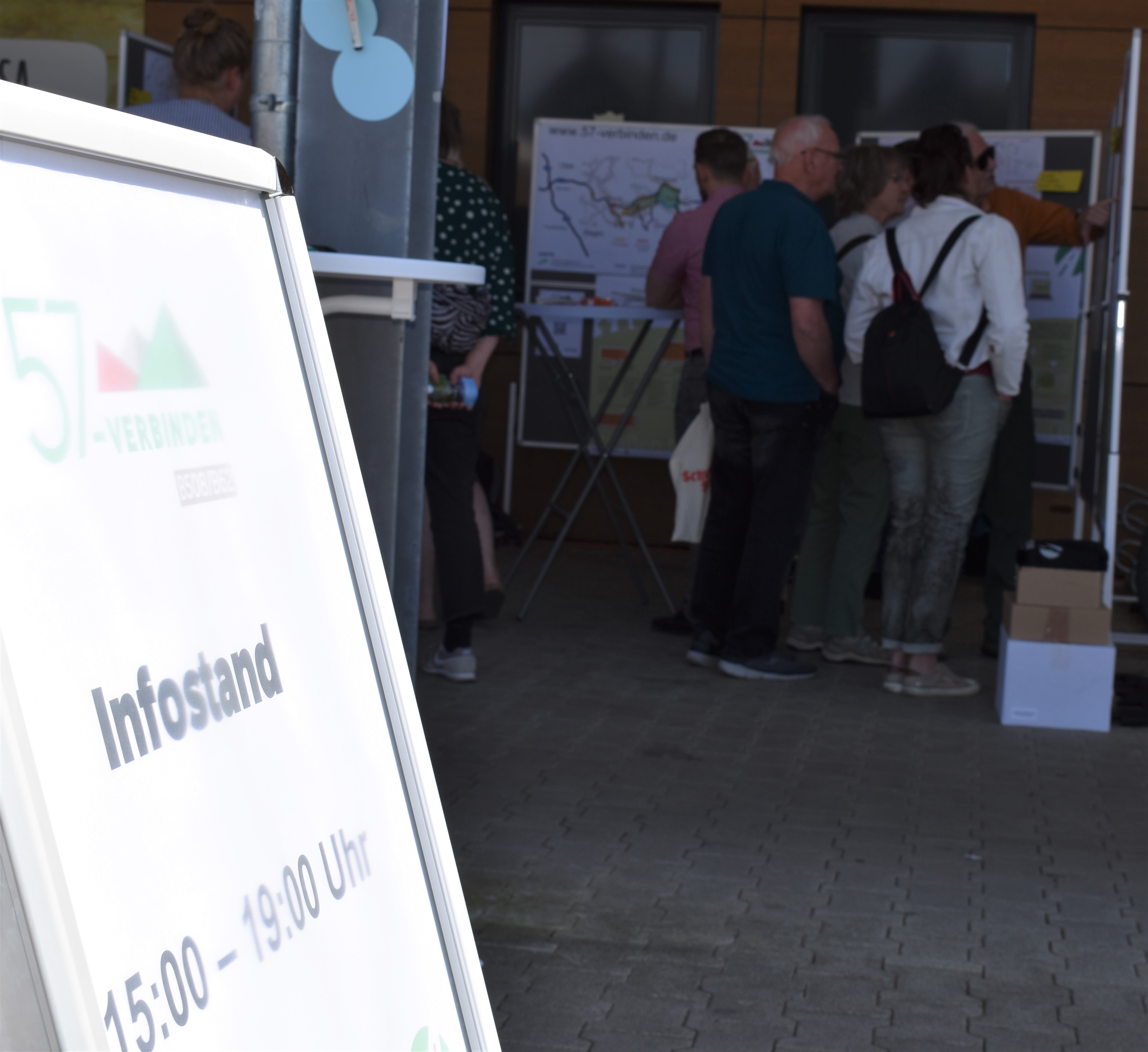 Im Vordergrund steht ein Austeller, der über den Infostand informiert. Im Hintergrund sind mehrere Personen vor Planungswänden zu sehen.