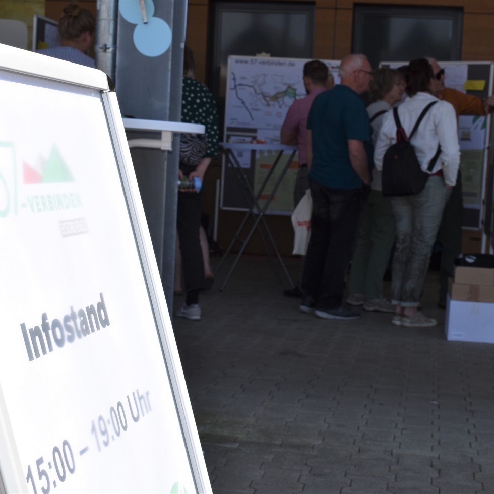 Im Vordergrund steht ein Austeller, der über den Infostand informiert. Im Hintergrund sind mehrere Personen vor Planungswänden zu sehen.